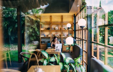 Yaratıcı kütüphanedeki ahşap masada oturan ve yaz tatillerinde laptopta ödev yapan konsantre genç etnik kız öğrenci.