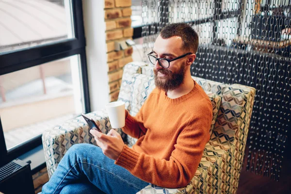 快乐的年轻人一边喝着热饮 一边坐在咖啡店的扶手椅上 一边在手机上读短信 — 图库照片