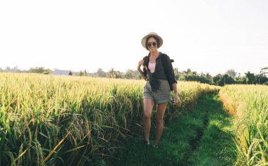 Kırsal kesimde yaz mevsiminde hasatla gülümseyen neşeli kadın turist, güneş gözlüklü mutlu bir Kafkasyalı kadın, uluslararası keşif sırasında kendini kaygısız hissederek saha yolunda yürüyor.