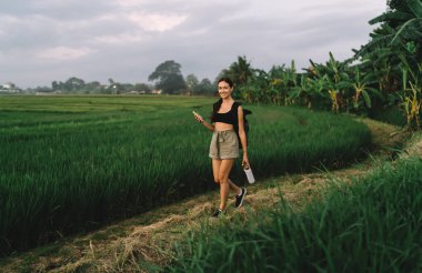 Bali 'de elinde akıllı telefonuyla pirinç tarlasında yürüyen gündelik kıyafet ve sırt çantasıyla gezen mutlu kadın yolcuların tüm vücudu.