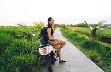 Bali 'deki pirinç tarlasında motosiklet üstünde oturan ve başka tarafa bakan, günlük kıyafetlerle gezen mutlu kadın turistin yan görüntüsü.