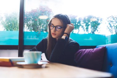 Siyah elbiseli, genç, düşünceli bir kadın serbest yazar masaya oturur, elini uzatır ve kafede çalışırken not defterine not alır.