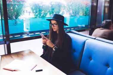 Kafeteryadaki masada oturan ve dinlenirken sosyal ağları kontrol eden şapkalı ve gözlüklü mutlu bir kadından.