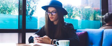 Şapkalı, gözlüklü, kafasında bir bardak sıcak içecekle masada oturan ve tatilde akıllı telefonlarla sörf yapan geniş açılı bir kadın.