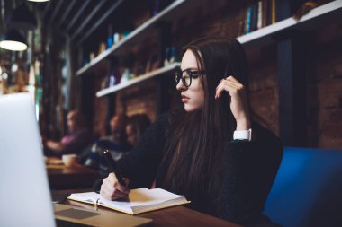 Ciddi kadın uzaktan çalışan sıradan giysiler içinde masaya oturmuş saçlara dokunurken ve not defterine notlar yazarken gözlerini kaçırıyor.