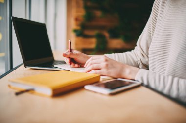Modern ofisteki iş günlerinde not defterine notlar yazarken bilgisayarlı ve akıllı telefonlu bir masada oturan ekin sahibinin yan görüntüsü