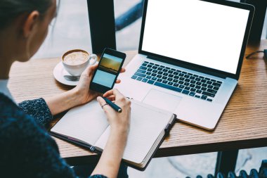 Dizüstü bilgisayarı ve kahve bardağıyla masada oturan serbest çalışan, akıllı telefondaki resimlerin tarifini not defterine yazan kadın.
