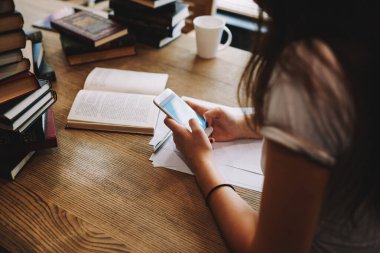 Kitap ve kupalarla ahşap masada otururken akıllı telefon çeviren tanınmayan bir kız öğrencinin yüksek açılı görüntüsü
