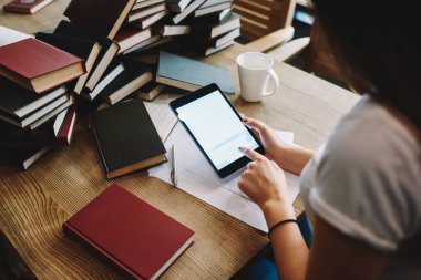 Tablet üzerinde internet sörfü yapan kadın kütüphanecinin yüksek açılı görüntüsü. Masada gazete ve kupalarla otururken.