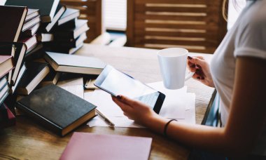 Tablet kullanarak kitap okuyan ve ödevlerini yaparken sıcak içecek içen kız öğrencilerin yan görüntüsü.