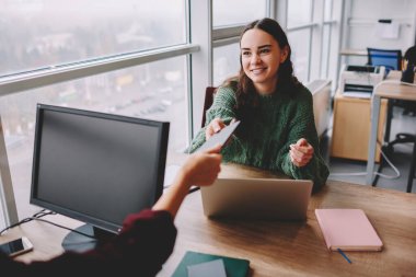 Yüksek açılı, dişlek tebessümlü, tahta masada dizüstü bilgisayarla oturan, iş arkadaşına tablet veren, hafif çalışma alanında birlikte çalışan genç bir kadın.
