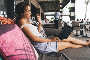 Kanepede dizüstü bilgisayarıyla oturan ve video konuşma sırasında ekrana bakarken el sallayan neşeli kadının yan görüntüsü