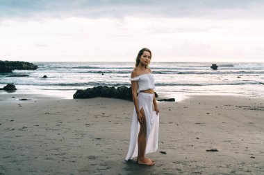 Gün batımında kumlu sahilde dalgalanan okyanusun kenarında beyaz elbiseli, çıplak ayaklı, genç ve güzel bir bayan gezgin.