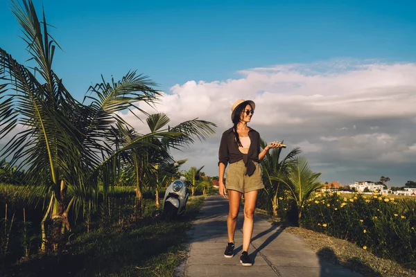 暑假期间 带着智能手机的快乐女性游客走在阳光灿烂的路上 探索热带乡村环境 带着手机漫步在博客上 — 图库照片