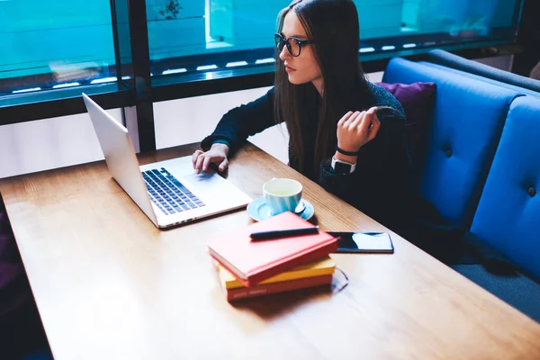 Uzaktan kumandalı, günlük kıyafetlerle, uzun saçlı, tahta masada bir fincan çayla oturan ve kafeteryada dizüstü bilgisayarla çalışan bir kadın.