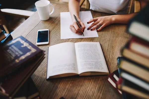 Yukarıdan gelen yüzü olmayan kadın boş bardak ve akıllı telefonla masada oturmuş kitap okuyor ve kağıda notlar yazıyor.