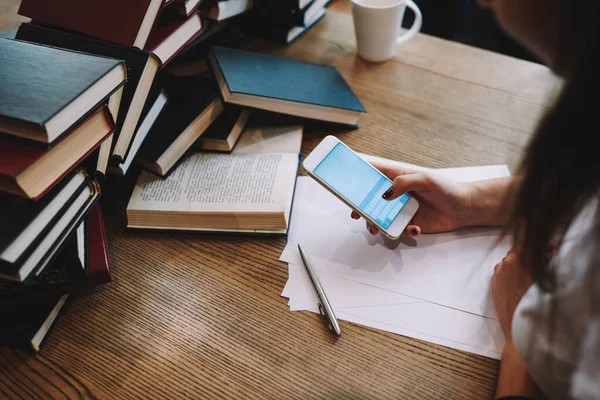 Kadın serbest çalışanın yaratıcı projede çalışırken cep telefonuyla mesaj göndermesi ahşap masada kağıt kalem ve kitaplarla oturması.