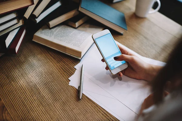 Proje için makale yazarken akıllı telefonu arayan yüksek açılı bir bayan öğrenci. Elinde kağıt defterleri ve kupalarla ahşap masada oturuyor.