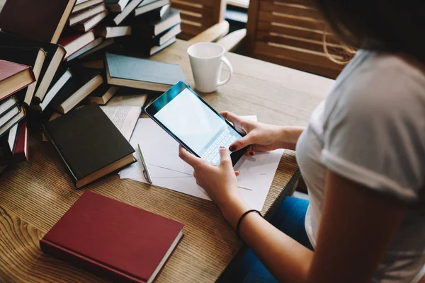 Yeni kreatif projede çalışırken tablet üzerinde internette sörf yapan genç bir kadın kitap ve kupalarla masada oturuyor.