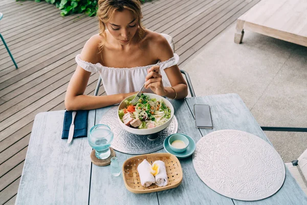 Beyaz elbiseli yüksek açılı, çıplak omuzlu, tahta masada oturan taze salata ve bir bardak suyla dolu bir kadın.