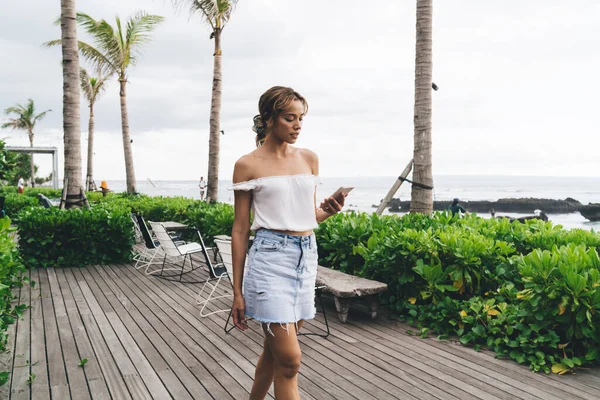 Düşünceli bir kadın, günlük giysiler içinde deniz kenarındaki ahşap terasta yürüyor ve ekranda cep telefonuyla mesajlaşıyor.