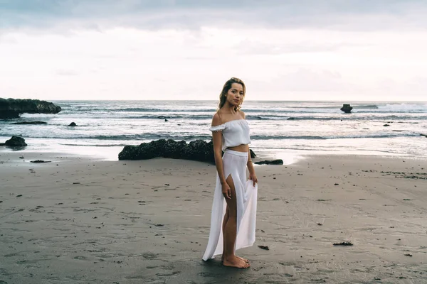 Gün batımında kumlu sahilde dalgalanan okyanusun kenarında beyaz elbiseli, çıplak ayaklı, genç ve güzel bir bayan gezgin.