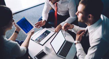 Yukarıdan gelen meslektaşlar dizüstü bilgisayar ve diyagramlı pano ile masada oturuyor ve ofiste projenin detaylarını tartışırken tablet kullanıyorlar