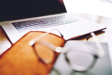 Gündüz vakti deri kapaklı ve gözlüklü, ahşap masadaki siyah ekranlı modern dizüstü bilgisayara yakın çekim.