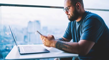 Hafif çalışma alanında yeni bir proje üzerinde çalışırken cep telefonu kullanarak masada oturan sakallı adamın yan görüntüsü