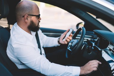Klas kıyafetli sakallı bir erkek sürücünün rahat bir arabada oturup trafik sırasında akıllı telefondan mesaj okuması.