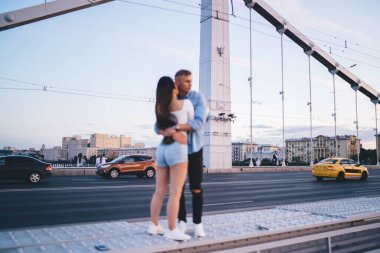 Günbatımında gökyüzüne karşı yolun kenarında duran ve kız arkadaşına sarılan gündelik elbiseli genç bir adam.