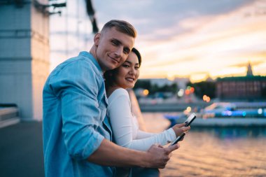 Günbatımında gökyüzüne karşı köprüde durup kameraya bakarken modern aletleri kullanan pozitif genç çiftin yan görüntüsü