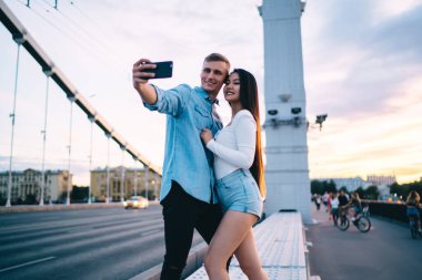 Gülümseyen genç erkek, gün batımının arka planında gezinirken uzun saçlı kız arkadaşıyla fotoğraf çekmek için akıllı telefon kullanıyor.