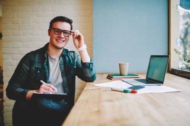 Genç bir erkek, dişlek bir gülümsemeyle masada dizüstü bilgisayarla oturmuş iş sırasında gözlüğe dokunan kameraya bakıyor.