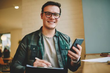 Genç bir erkek, dişlek gülüşü ve gözlükleriyle kameraya bakıyor ahşap masada oturuyor, akıllı telefonuyla kafede vakit geçiriyor.