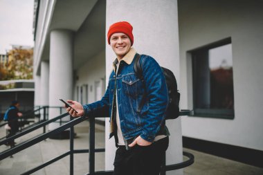 Yan görüntüde, cebinde cebi olan, parmaklıklara yaslanmış, binanın yanında duran ve akıllı telefonları karıştıran, sokakta boş zaman geçiren genç bir erkek görülüyor.