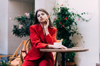 Kırmızı üniformalı genç bir bayan açık kafede oturmuş çenesine dokunuyor ve düşünceli bir şekilde kameraya bakıyor.