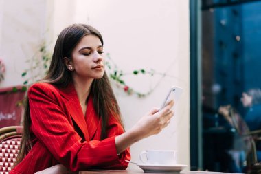 Kırmızı elbiseli bir kadın, gün ışığında beyaz ve cam duvarlı rahat bir cadde kafesinde otururken akıllı telefonuyla internette sörf yapıyor.