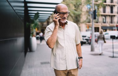 Resmi kıyafetler ve gözlük takmış, kaldırımda yürürken telefonla konuşan ve gözlerini kaçıran son sınıf sakallı, kel bir erkek.