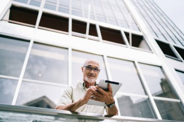 Klasik gözlüklü yetişkin erkek turistin portresi modern dokunma yastığını elinde tutuyor ve şehir merkezini keşfetmek için solo yolculuk sırasında kameraya bakıyor, dijital tabletli Kafkas blogcu