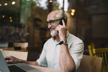 Optik gözlüklü neşeli yaşlı adam göz düzeltmesi için laptopun başında akıllı telefon sohbetinin keyfini çıkarıyor başarılı erkek serbest çalışan 60 'lı yaşlarda video izleyip gülümsüyor.
