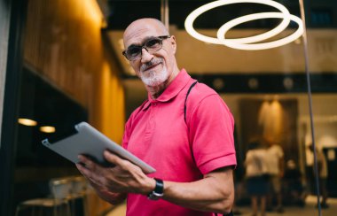 Gözlüklü, gözlüklü ve tabletli gülümseyen bir erkek gezginin dışarıdaki haritada kameraya ve tarayıcı uygulamasına baktığı düşük açı.