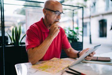 Kel kafalı yaşlı sakallı adam tablet ve seyahat haritasını izliyor. Masadaki kafede oturuyor.