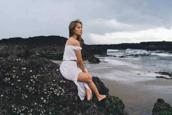 Beyaz elbiseli, çıplak ayaklı, ince bir kadın kayalıklarda oturuyor ve tatilin tadını çıkarırken dikkatlice başka tarafa bakıyor.