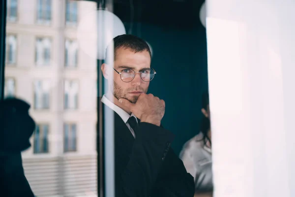 現代オフィスのガラスの壁の近くに立つエレガントな正式なスーツ姿の若いビジネスマンとカメラを見ながらあごに触れる — ストック写真