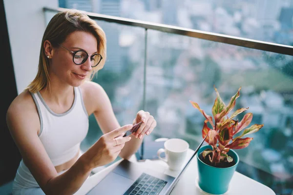Masada dizüstü bilgisayarla oturan ve balkonda içki içen genç kadın kartviziti yüksek açılı.