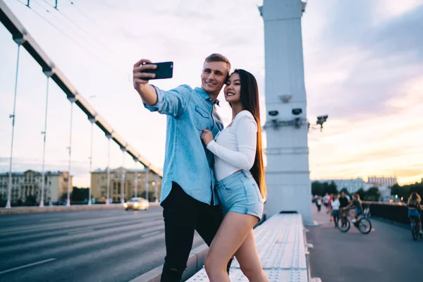 Gülümseyen genç erkek, gün batımının arka planında gezinirken uzun saçlı kız arkadaşıyla fotoğraf çekmek için akıllı telefon kullanıyor.