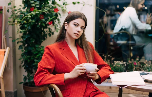 おしゃれなスーツ姿のシリアスな若い女性が昼間はストリートカフェで熱い飲み物を飲みながら丸テーブルに座っている — ストック写真