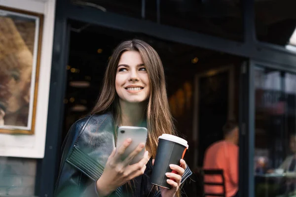 快乐的微笑女士 留着一头棕色的长发 头戴便服 手握咖啡和手机 站在城市街道上 在模糊的咖啡馆和笑声的背景下 — 图库照片