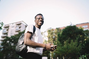 Gülümseyen koyu tenli erkek gezginin yarım boy portresi akıllı telefon ile 4G internete bağlanarak geziyor, gezmek için, neşeli Afro-Amerikalı adam sokaklarda gezinme uygulamasından memnun.